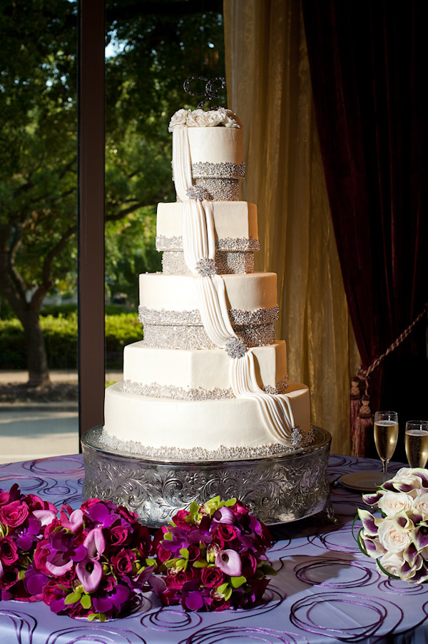 White five tiered wedding cake with silver beaded borders and a ivory drape with silver beaded brooches on each layer - cake is on vintage style cake stand with dark purple, lavender, and  pink floral decor surrounding - photo by Houston based wedding photographer Adam Nyholt
